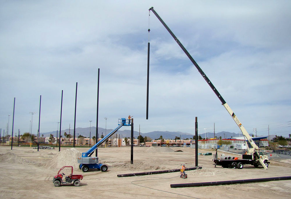 Construction site with cranes lifting and installing tall Coastal steel poles. Workers operate machinery on a flat, dirt terrain under a cloudy sky with distant buildings and mountains visible.
