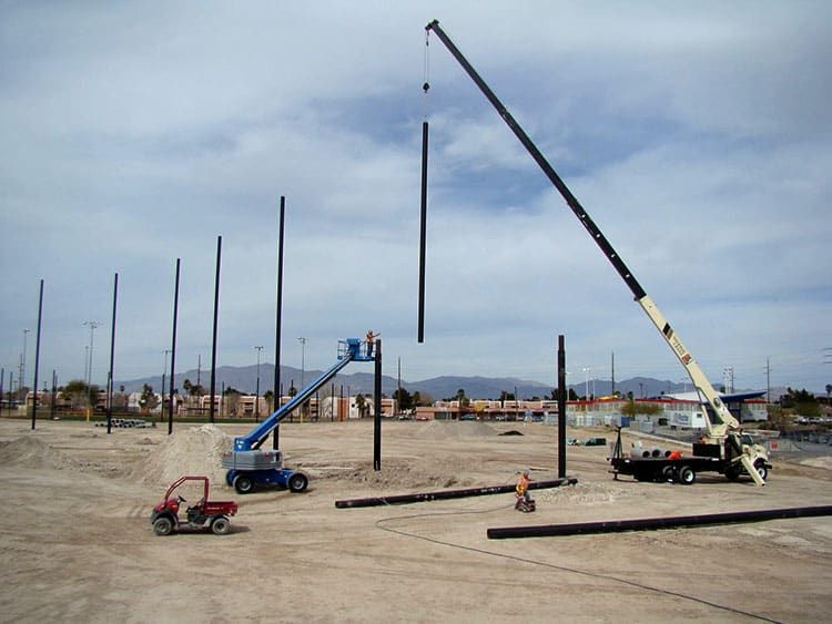 A construction site with cranes lifting Coastal&rsquo;s steel poles into place, workers in hard hats, and various machinery on a dirt surface under a cloudy sky.