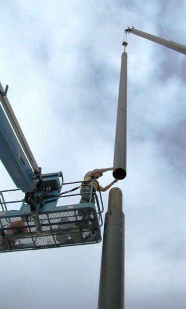 Looking up at a worker in a lift installing a tall Coastal Netting steel pole using a crane under a cloudy sky.