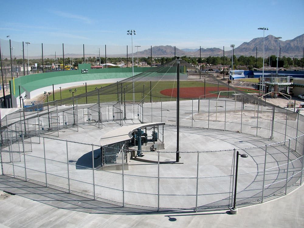 A baseball batting cage surrounded by a chain-link fence, with adjacent baseball fields and distant mountains under a clear sky.