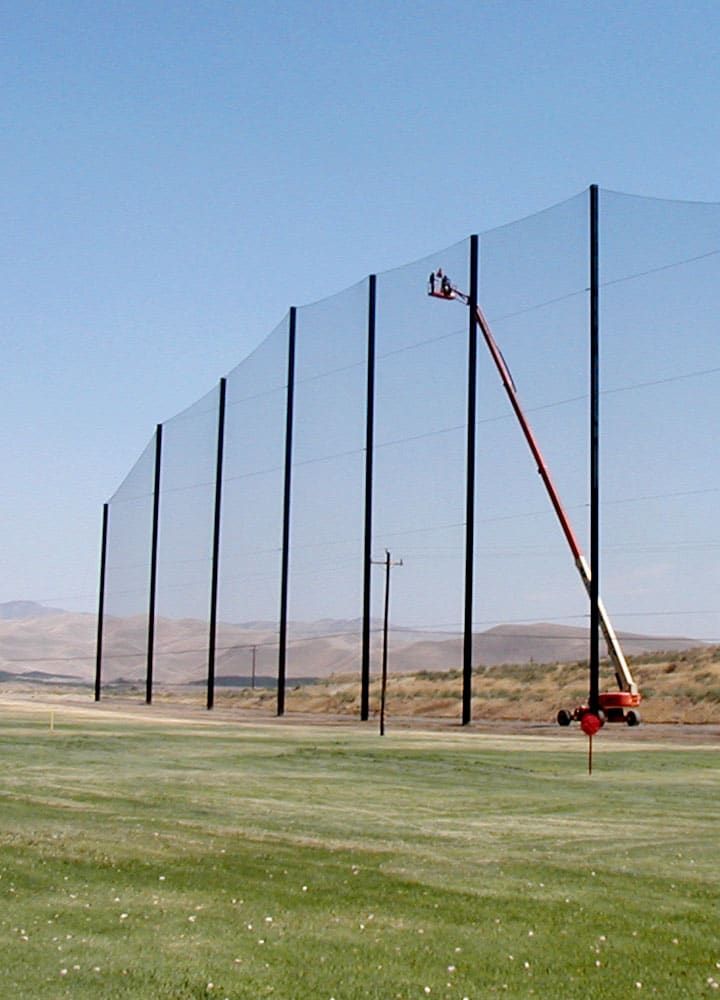 A worker high up on a cherry picker during installation of a tall netting system at the edge of a driving range. There are mountains in the background under a clear blue sky.