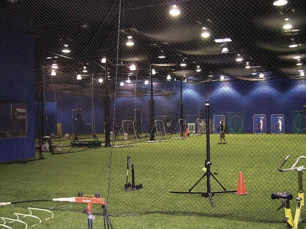 Indoor sports facility with turf field, sports equipment including targets and training gear, surrounded by netting. Floodlights illuminate the space; a person walks in the distance.