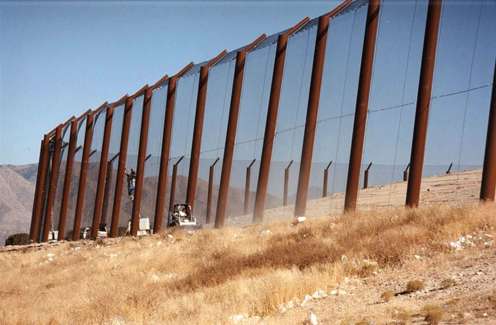 A tall stationary litter fence runs along a dry, grassy landscape with a rugged terrain in the background. A person is seen on a lift working on the fence, while a bobcat vehicle drive nearby.