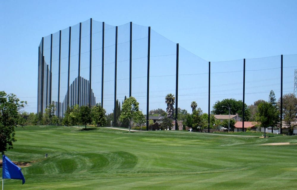 Golf course with tall black netting poles in the background, blue flag on a foreground green. Trees and houses are visible beyond the netting. Clear blue sky.